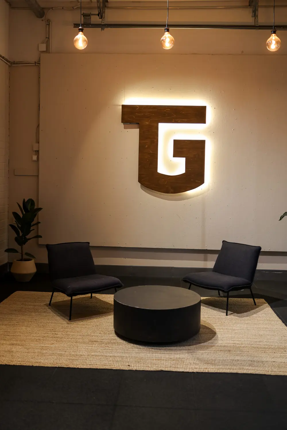 Salin aula: valaistu puinen TG-logo seinällä, kaksi nojatuolia, pyöreä pöytä ja viherkasvi.