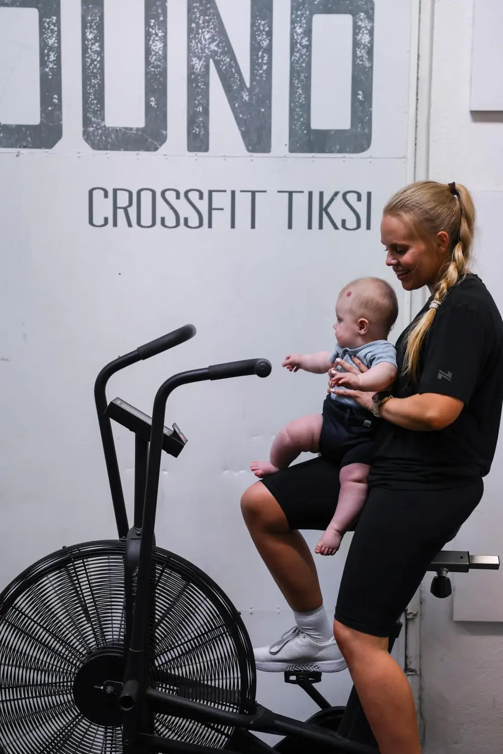 Äiti istuu kuntopyörän satulassa vauva sylissään salin seinän Training Ground -tekstin edessä.