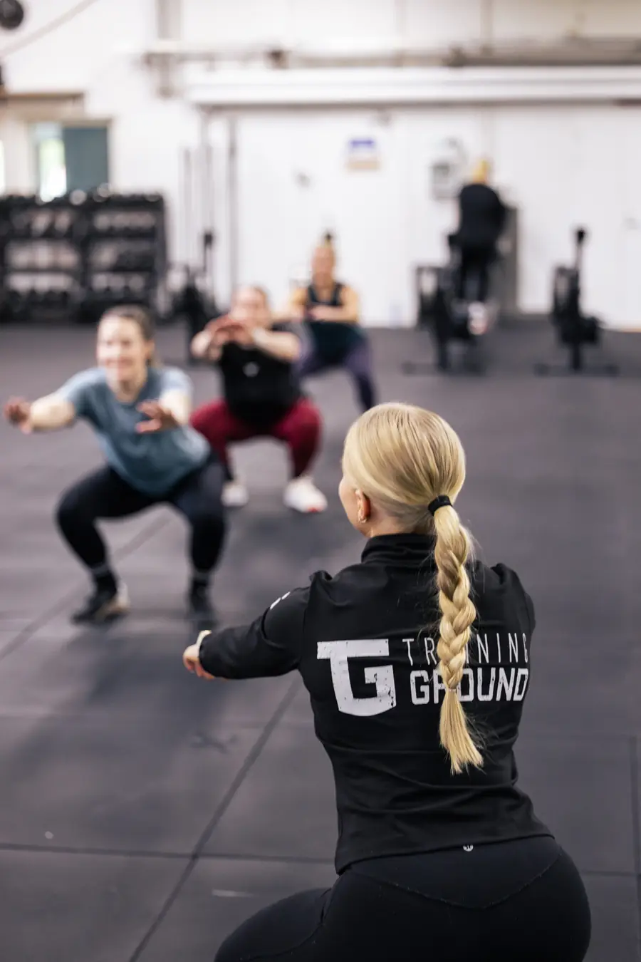 Valmentaja Training Ground -takissa ohjaa kyykkyjä tekevää ryhmää.